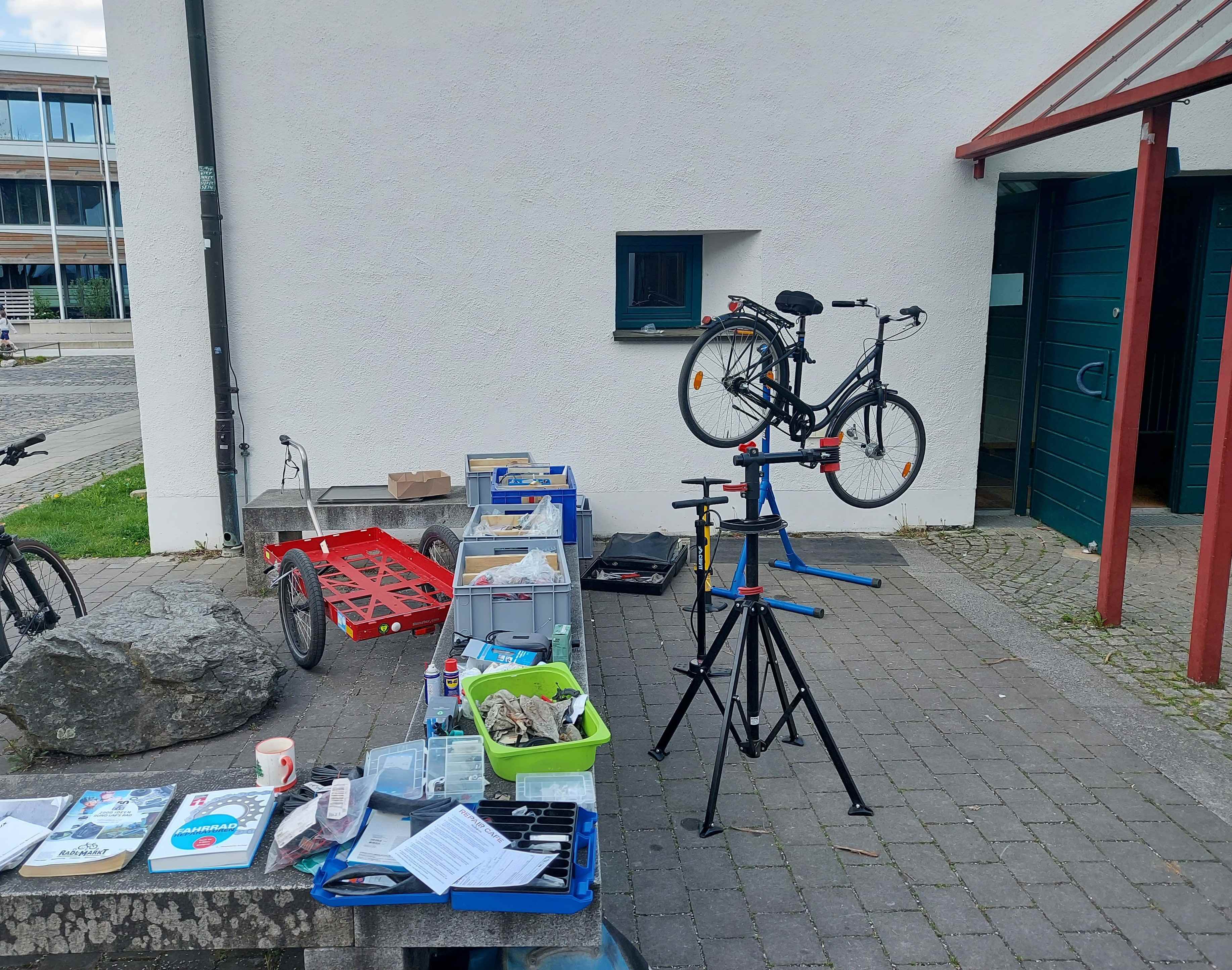 Alt-Text: Ein provisorischer Fahrrad-Reparaturstand im Freien vor einem weißen Gebäude. Auf einer Steinbank liegen Werkzeuge, Kisten mit Ersatzteilen und Infomaterial. Ein schwarzes Fahrrad ist in einem Montageständer aufgebockt, daneben stehen ein roter Fahrradanhänger und eine Luftpumpe bereit.