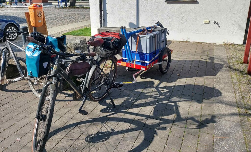 Alt-Text: Ein schwarzes Tourenfahrrad mit blauen Packtaschen steht auf einem gepflasterten Platz. Ein zweites Fahrrad zieht einen roten Lastenanhänger, der hoch mit grauen und blauen Transportkisten beladen und mit Spanngurten gesichert ist. Die Szene ist bei sonnigem Wetter im Freien aufgenommen.